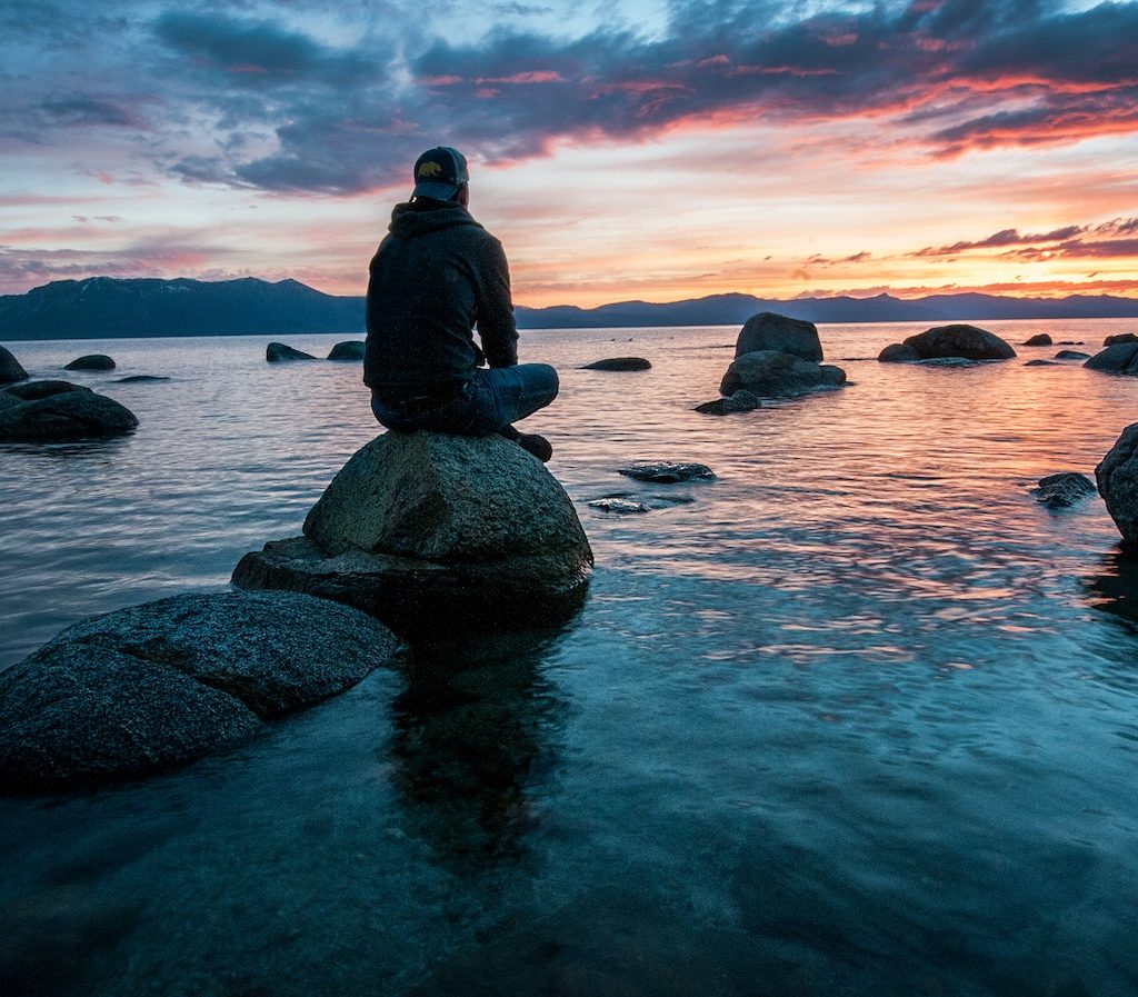 junger Mann sitzt alleine auf einem Stein an einem See und schaut in den Sonnenuntergang - Einsamkeit
