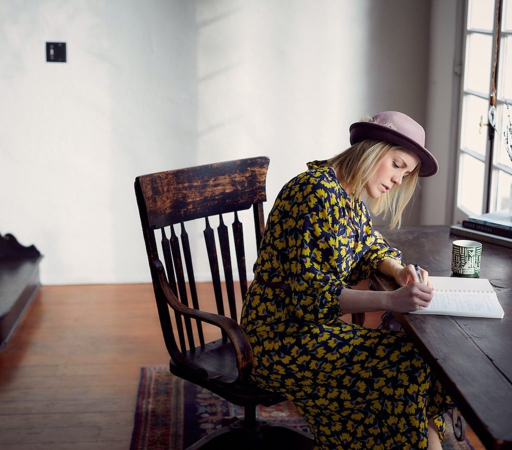 junge blonde Frau mit Hut sitzt am Schreibtisch und schreibt in ihr Tagebuch Journal