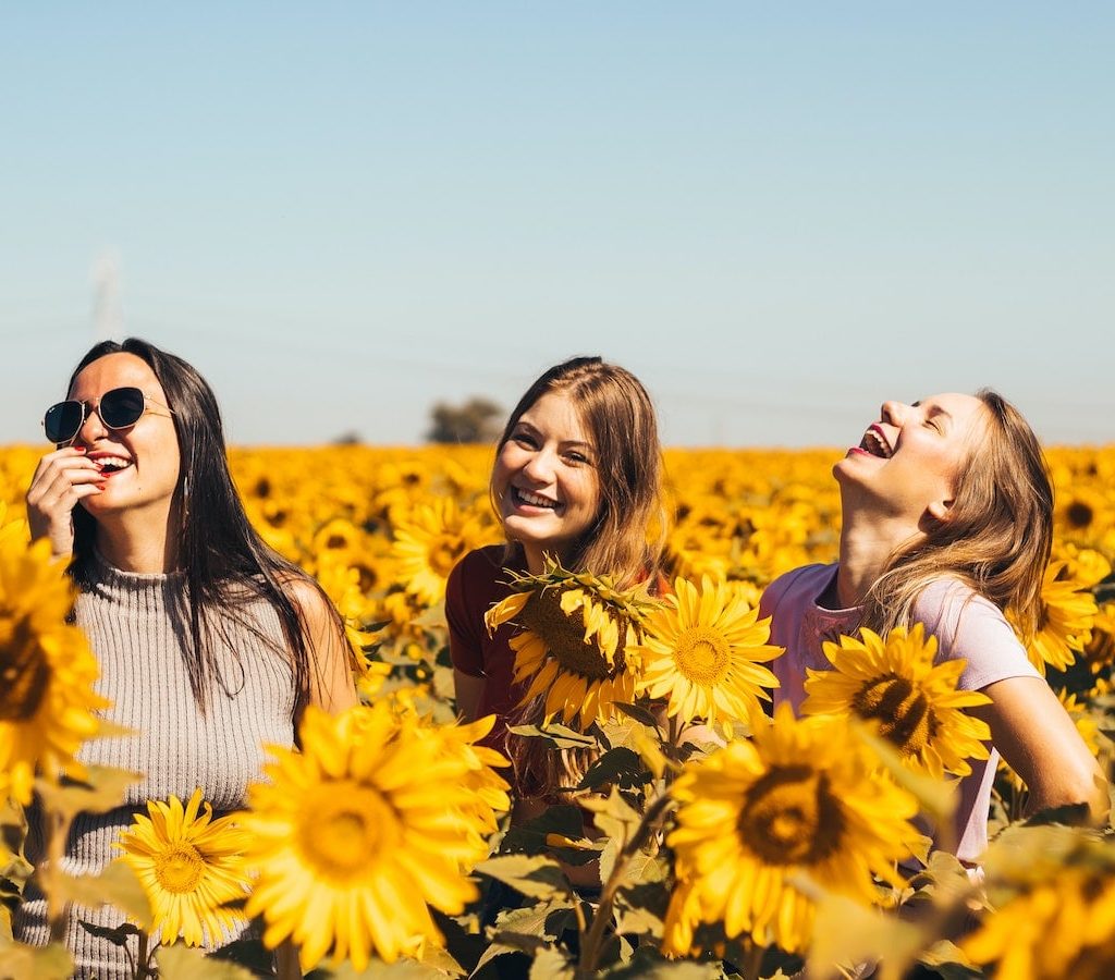 drei junge Frauen lachen gemeinsam in einem gelben Sonnenblumenfeld - mit Freunden Einsamkeit überwinden