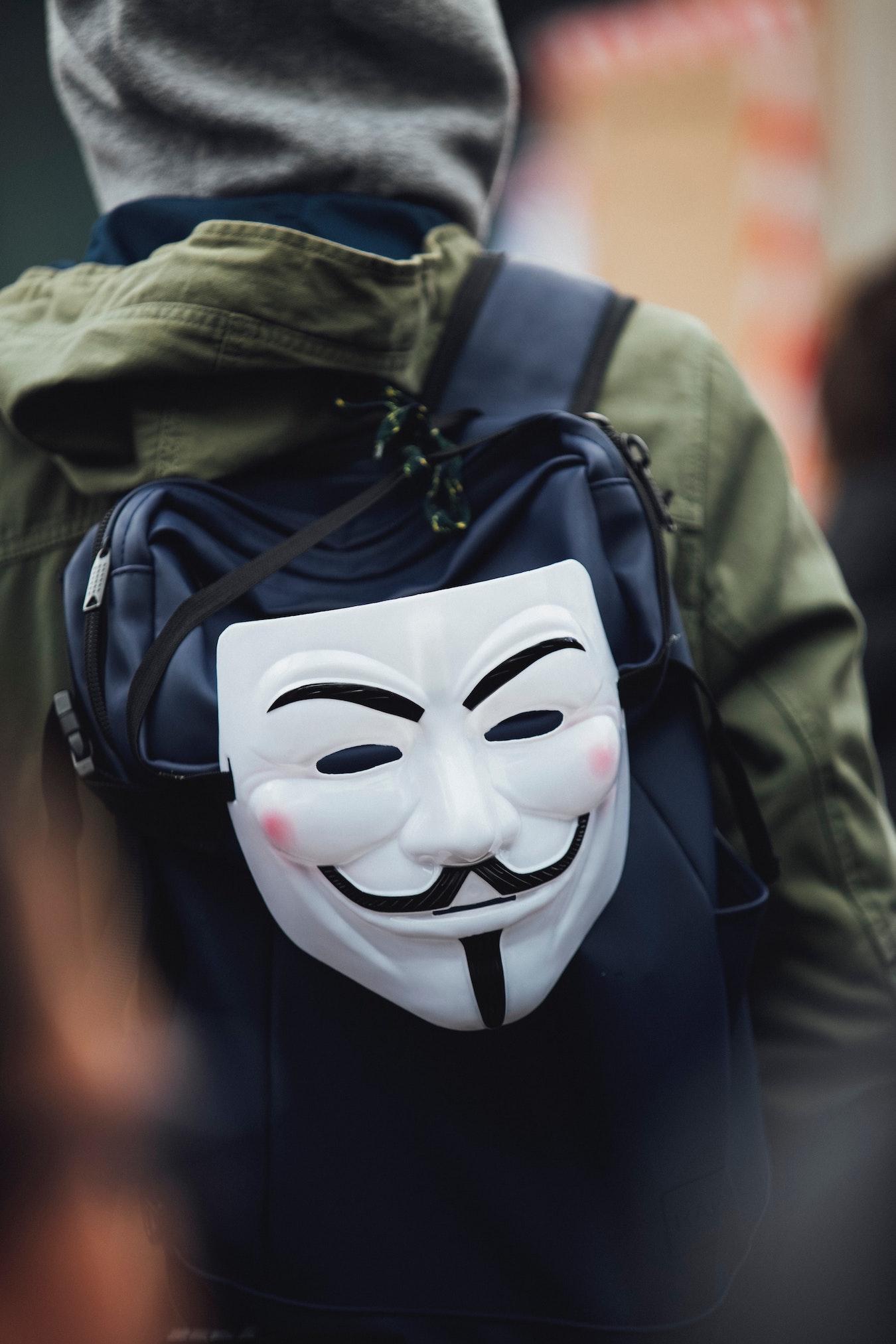 Rücken einer Person mit Rucksack, an dem eine Anonymus Maske hängt