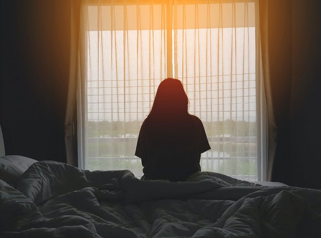 junge Frau sitzt einsam auf ihrem Bett und schaut aus dem Fenster raus in die Ferne - Einsamkeit überwinden