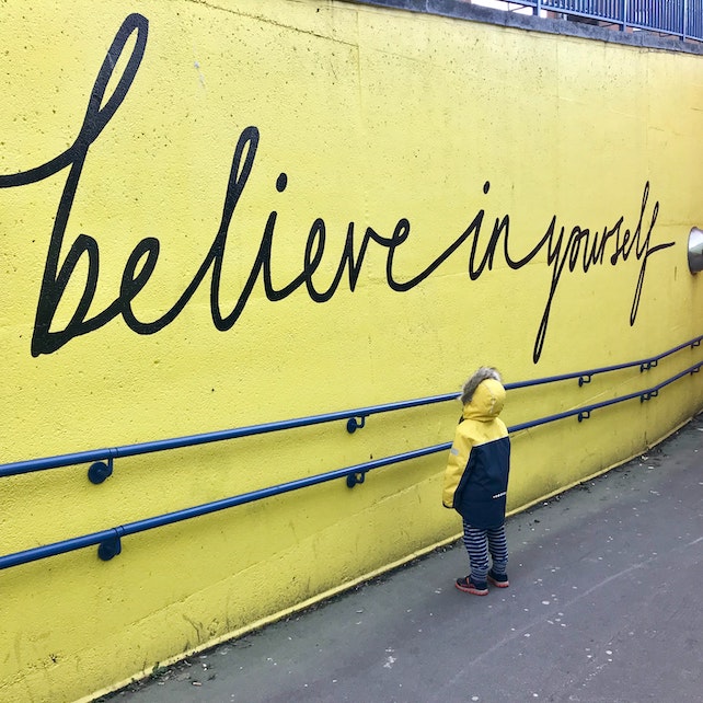 Glaube an dich selbst kleines Kind steht vor einer gelben Wand mit der Aufschrift believe in yourself - englisch für glaube an dich selbst