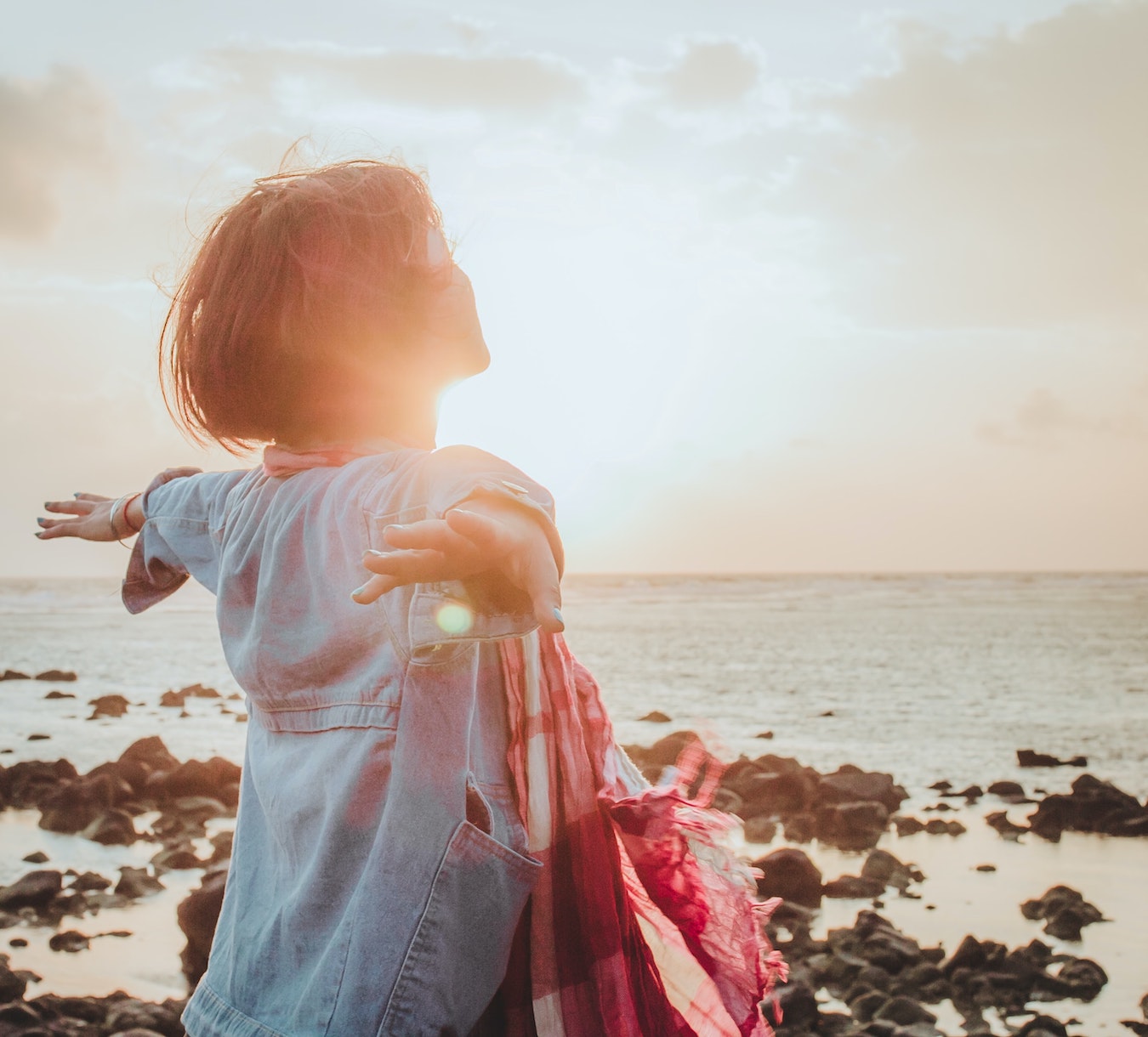 junge Frau schaut auf das Meer in die Sonne und hält ihre Arme weit offen Freiheit