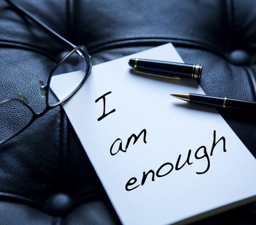 ein Zettel mit der Aufschrift "i am enough", zu Deutsch "ich bin genug" liegt neben einer Lesebrille auf einer schwarzen Ledercouch